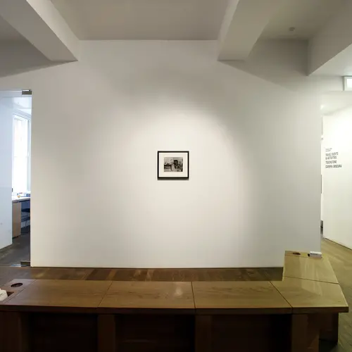 Gallery room with small framed black and white photograph and a dog-legged bench in front. 
