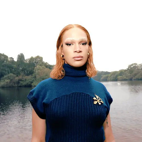 A person dressed in a cropped blue jumper stands in front of a lake on a overcast day.