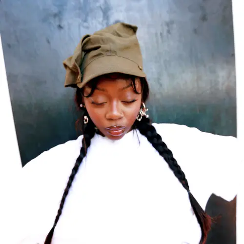 A young woman with long braided hair and a grey hat on stands with her arms out.