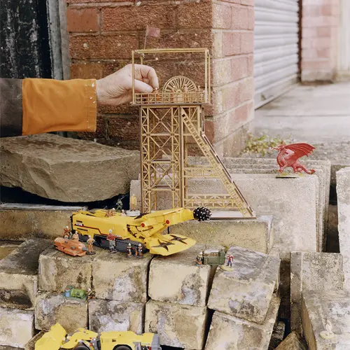 A child's hand reaches into the frame to hold a small figurine which is being placed inside a miniature wooden mill. The mill is stood on top of a pile of bricks which has a few toy tractors, diggers and bulldozers placed around.