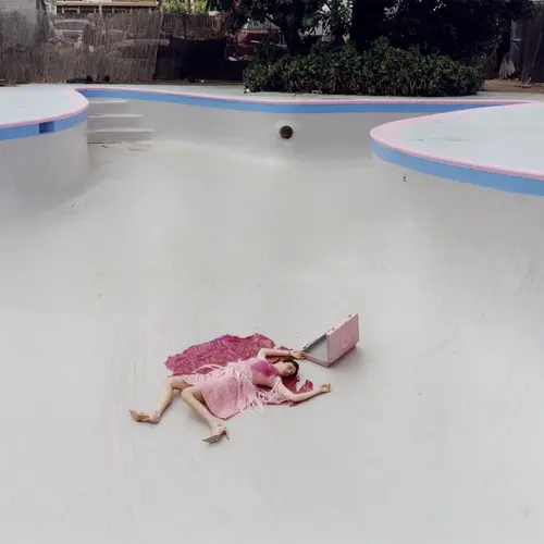 A woman in a pink floaty dress lays sprawled at the bottom of an empty pool.