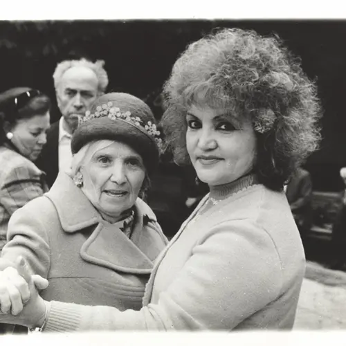 Black and white photograph of a woman  and an older woman holding hands as they dance together.  An older couple can be seen in the background