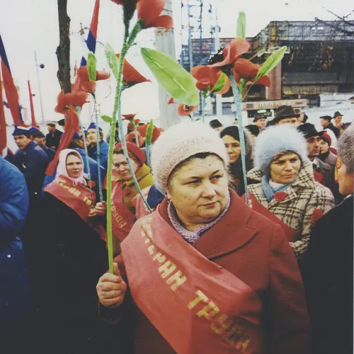 Colour photograph of a woman  standing amongst a crowd of people. She is wearing a red  sash across her body and is holding up a fake red flower.