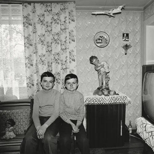 Black and whit eimage of two young Polish boys who are identical twins. They are sat on a chairs next to a side table with a ceramic figurine of a cherub on it.