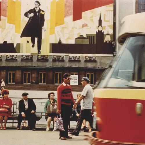 Colour photograph of people sat on a bench  with a large billboard of Vladimir Lenin behind. A tram can be  seen in the corner passing in front.