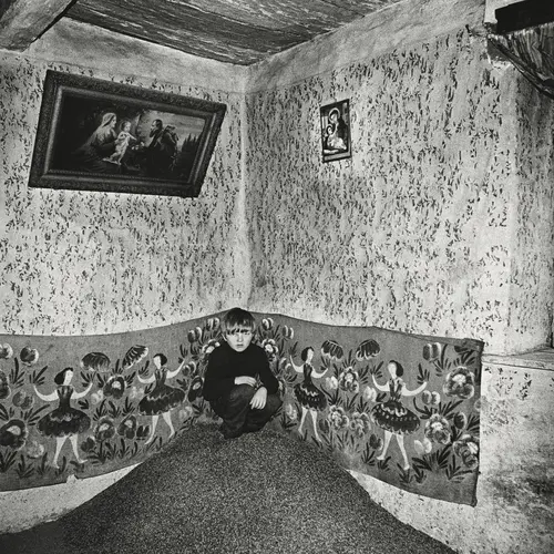 Black and white image of a young Polish boy crouched in a rustic room which has been carefully decorated with framed biblical paintings.