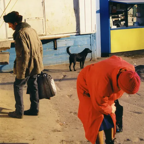 Colour photograph of an unidentifiable  person wearing a red coat ands red hat crouching down  and a man wearing a winter coat and hat holding luggage. A  black dog can be seen at the back of the photo.