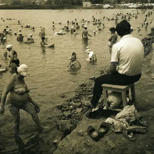  Sepia photograph of a people frolicking in a  body of water. Women and men can be seen in bathing suits  aside from one man who is captured sitting at the edge of  the bank on a stool in a white shirt and black trousers.
