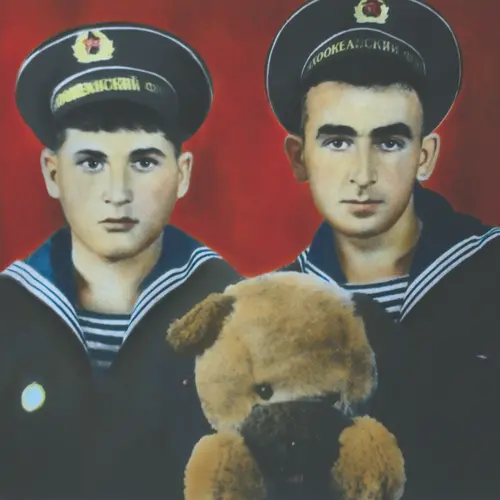 Colour photograph of two young men in  navy uniform in front of a red background with a soft toy  seen in front