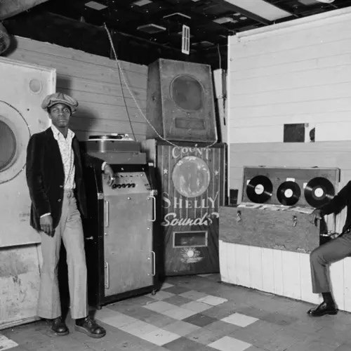 Two young black men sit in a basement club surrounded by sections of a sound system.