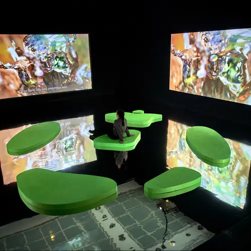 A woman watches a film being shown on two screens in a dark, reflective space, while seated on one of five green shapes 