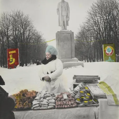  Colour photograph of a woman  standing in front of a monument selling food items. She  wears a white coat and a furry blue hat.