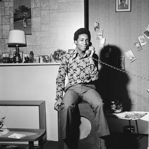 Black and white photograph of a young man sitting on a speaker with a telephone receiver held to his ear.
