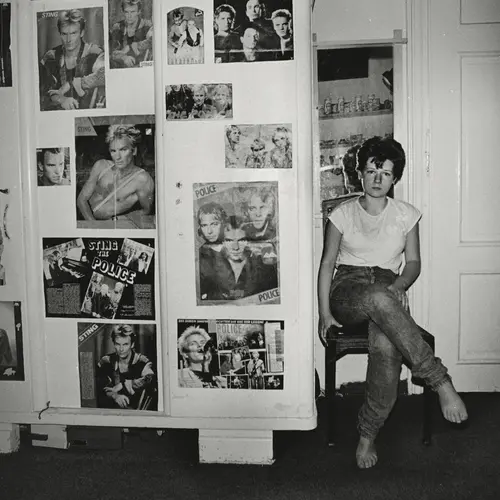Black and white image of a young Polish woman sat on a wooden chair next to a wall full of posters of posed singers and artists.