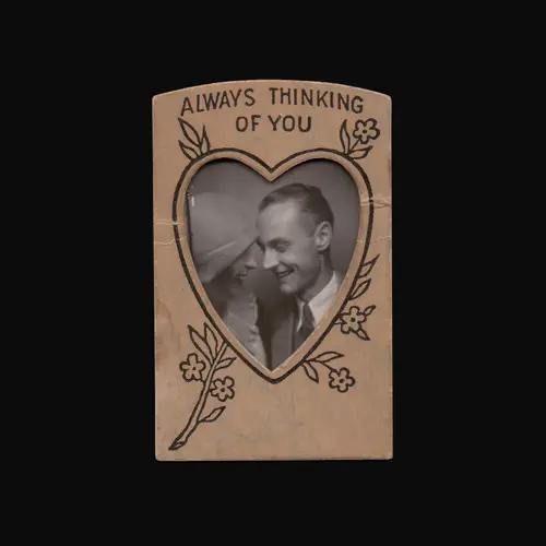 A scan of a vintage paper photoframe which is cut out into a heart shape and printed with 'Always Thinking of You'. It houses a black and white photograph of a couple smiling at one another.