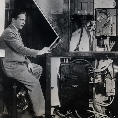 A black and white vintage photograph of man sat inside of a early photobooth.