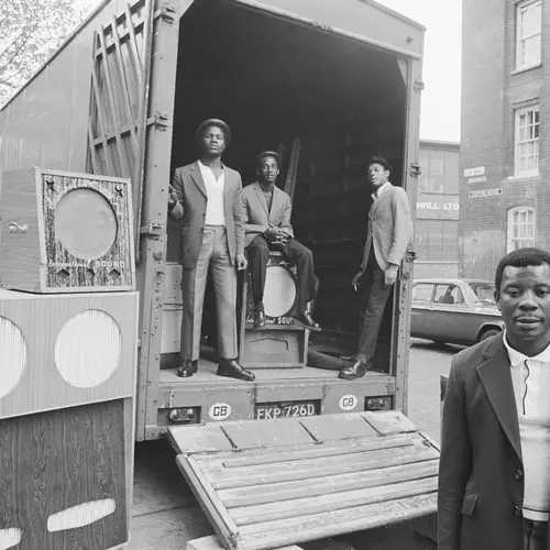 Black and white photograph of three men standing in the back of a truck parked next to a soundsystem. A man is also stood in front of the truck.