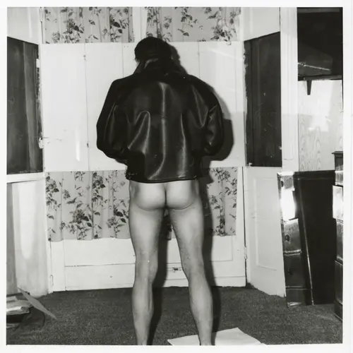 Image of a trouserless and pantless man standing in a living room wearing a leather jacket with his back to us.