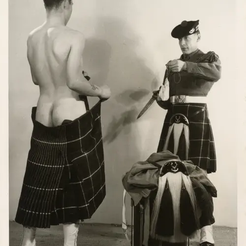 One man wearing a kilt skirt faces another man wearing a full kilt outfit, pointing a knife at the other