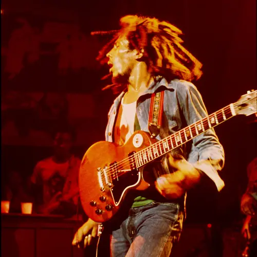Colour photograph of Bob Marley with a guitar slung across his body in a live performance.