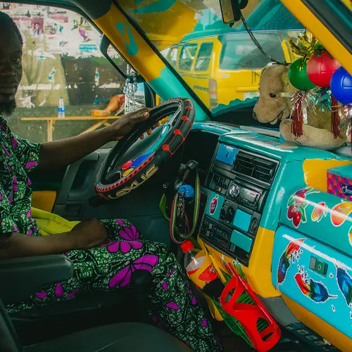 A close up of a driver sat behind the wheel of his vehicle. He wears a traditional, Nigerian patterned outfit. His dashboard is wrapped in turquoise vinyl and arranged with fruit stickers and small ornaments.