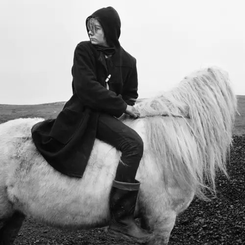 A black and white photo of a young person dressed in dark clothing riding a white horses with a shaggy, loose mane across a field of grass.