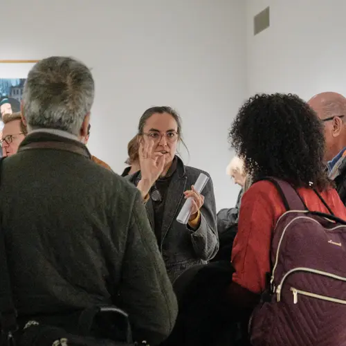 A woman wearing glasses with her hair tied back, leads a tour in an exhibition.