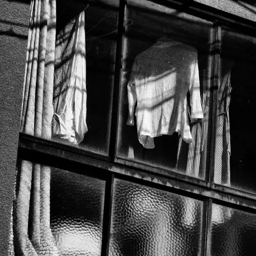 A black and white photograph of a window taken from an external perspective. There are items of clothing hung up to dry in view between the curtain drapes.