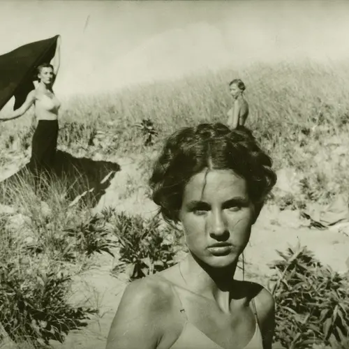 Black and white photograph of a woman staring directly at the camera. Three women can be seen standing in the distance.
