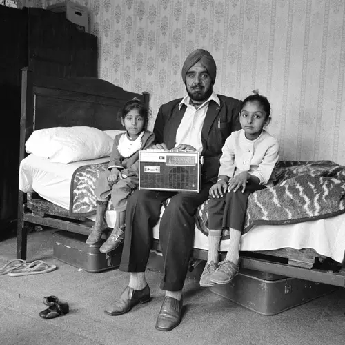 Black and white photograph of a man sitting on a bed with his two small daugters sat on either side of him. A radio set is perched on the man’s lap.
