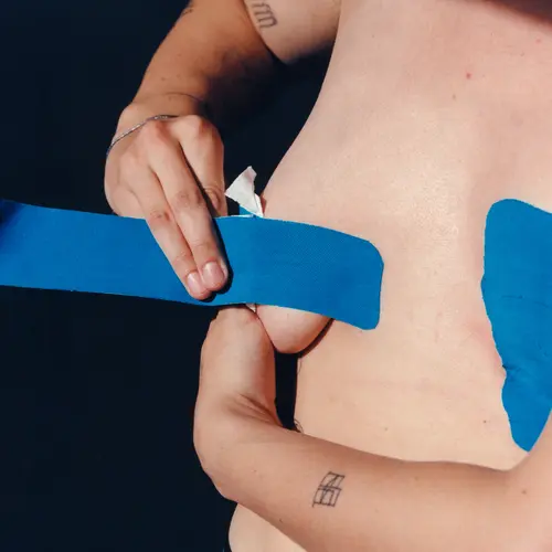 A close up of someone using blue body tape to bind their breasts.