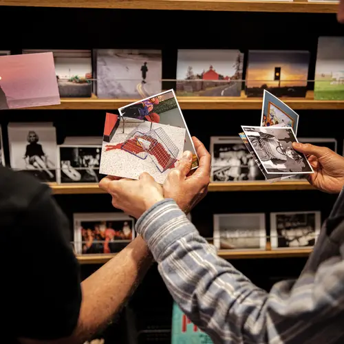 A close up shot of two sets of hands holding a selection of postcards in fron tof a shelf full of postcards on sale.