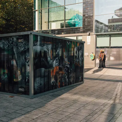 Install image from V1: Content Aware in which a large shipping crate installed with a camera is capturing images of itself in a mirrored window.