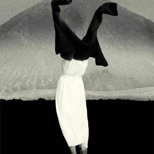 A black and white grainy image of a person dressed in white stood holding a jacket above their head as they slip it over their arms. Their face is covered by the jacket. A mountain is seen far away in the background.