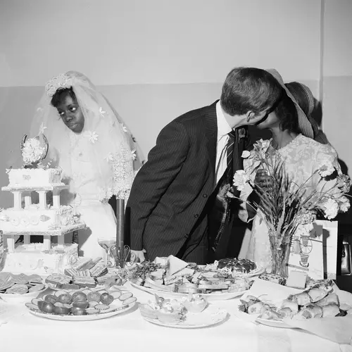 A young couple on their wedding day. The woman stares out over the cake with her wedding veil pulled back as her groom leans over to kiss the mother of the bride on the cheek.