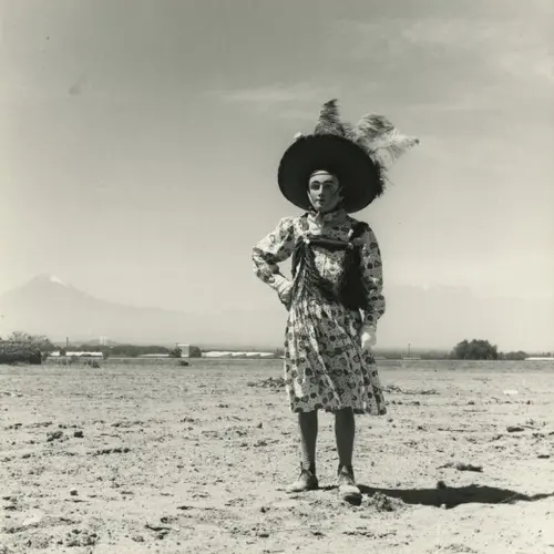 Black and white photograph of a masked individual wearing a dress and a fancy hat, standing in the desert