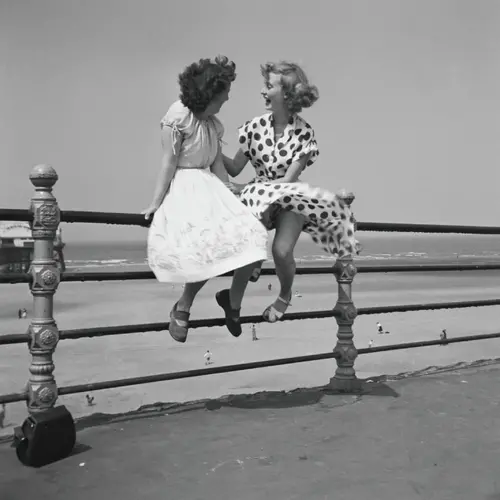 Black and white photograph of two women sat on a railing of a beach promenade