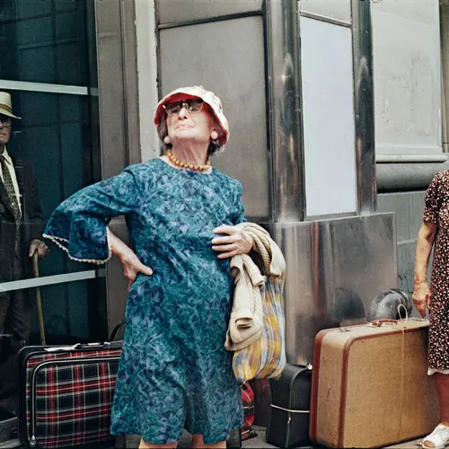 Two ladies have suitcases. One looks at the ground. One at the sky.