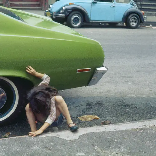 A young child squats under a car by a roadside. It is not clear whether she is urinating.