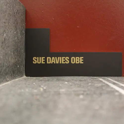 A black L shape with gold writing saying 'Sue Davies OBE'