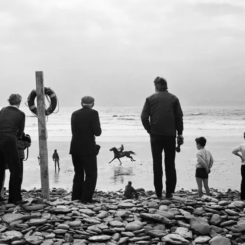 Black and white photograph showing a group of people standing on a rocky shore watching a person riding a horse.