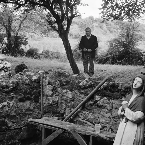 Black and white photograph depicting three people in an outdoor setting. Behind a low stone wall, one person sits holding a baby and another stands next a tree. A statue of Mary stands near a wooden bench in front of the wall. 