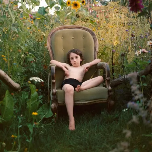 Colour photograph of a young person sitting in an armchair in a garden surrounded by plants and flowers