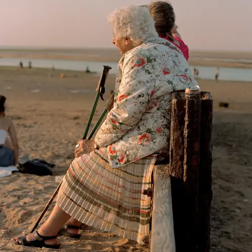 A colour image of an elder woman relaxing on a wooden support on the beach, looking out to the sea.