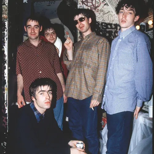 Colour photograph of Oasis posing for the camera. All of them are stood up with the exception of Liam Gallagher who is crouched down.