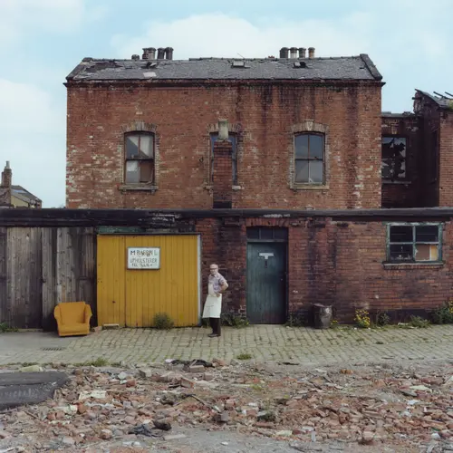 Colour photograph of the back of a house on a  street. An old man in an apron stands on the street. 