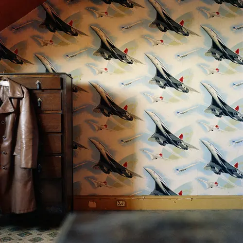  Colour photograph of a room with Concorde plane  wallpaper. To the left of the photo, a wooden chest of drawers can be seen with a brown leather coat hanging on it