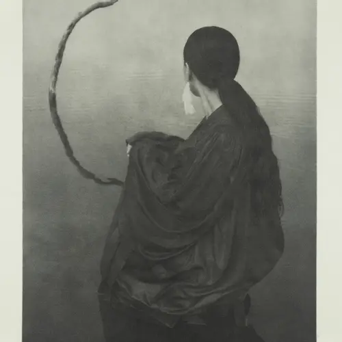 Black and white photograph of a person with long tied back hair, kneeling in a still, shallow body of water. They are facing away and looking at a curved branch protruding from the water.