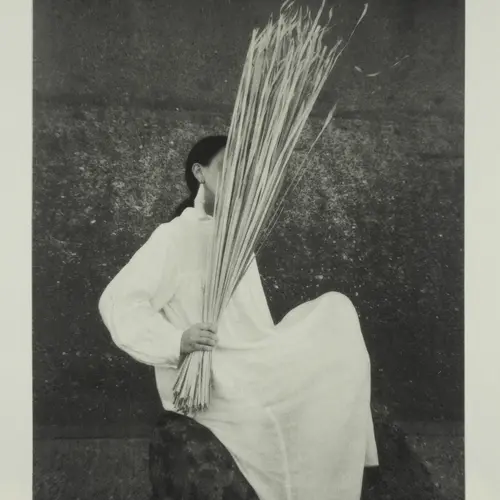 Black and white photograph of a person in light clothing holding a dense bundle of long, thin reeds vertically in front of their face.