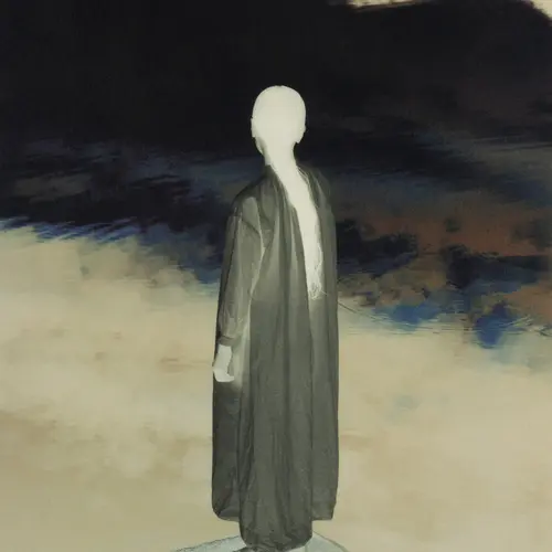 Inverted photograph of a person facing a dark, abstract background. The person wears a loose jacket to their ankles and has long hair tied back.